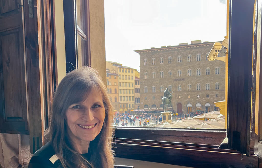 The author sitting beside a window looking onto the piazza from a bedroom in the Hotel In Piazza della Signoria.Copyright@2024 NancyRoberts 