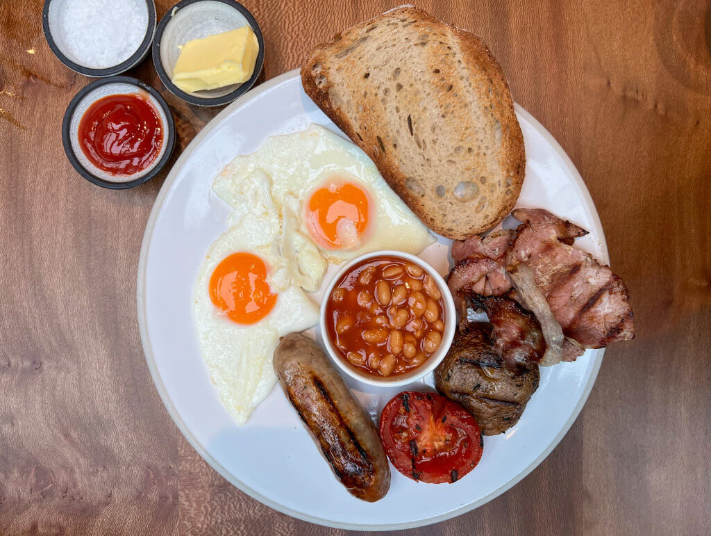 Typical London food: a full English breakfast on a white plate with toast, fried eggs, bacon, sausage, tomato, mushrooms, baked beans and condiments in dishes on the side. Copyright@2024NancyRoberts