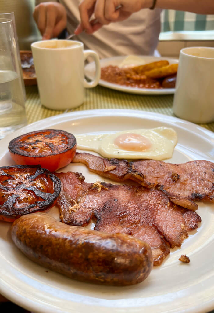 A full English breakfast in a greasy spoon cafe with mugs of tea beside plates of bacon, sausage, tomatoes and egg. Copyright@2024NancyRoberts