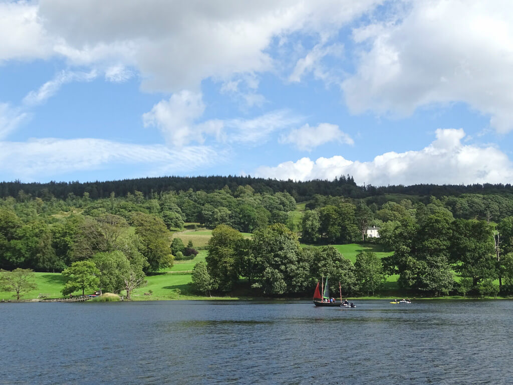 Coniston lake with vintage sailing boat with red sail and lush trees and lawns in background. Copyright@2025NancyRoberts