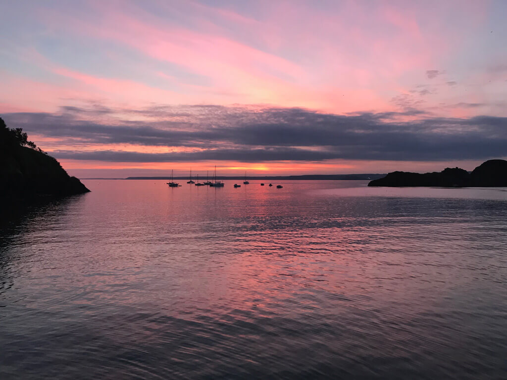 England countryside view of a beautiful pink and orange sunset spreading across a cove with small boats silhouetted in distance. Copyright@2025 mapandfamily.com 