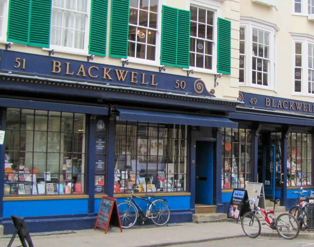 Bicycles parked outside traditional bookshop in Oxford with navy sign board and green shuttered windows above. Copyright@f2025 Nancy Roberts