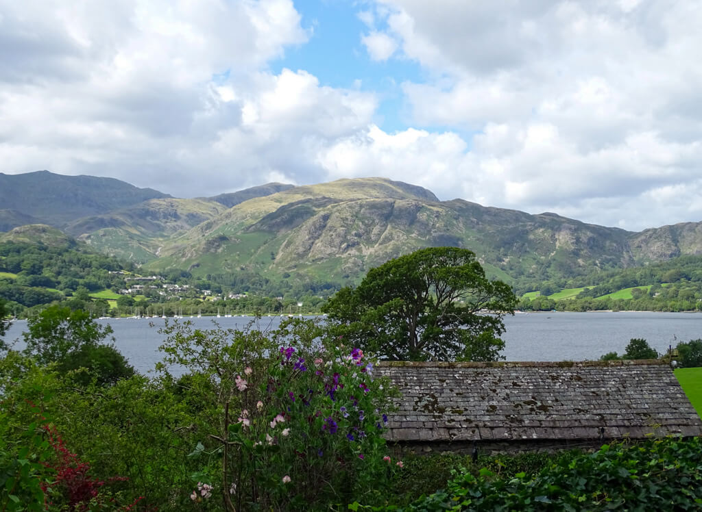 Lake district view of rolling hills behind a calm lake with small boats and wild flowers in foreground. Copyright@2025 NancyRoberts