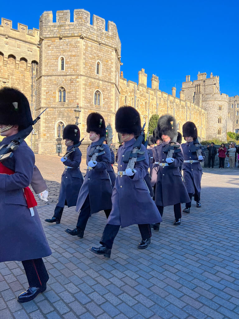 Soldiers in grey coats and busbys marching with guns on their shoulders in front of Windsor castle. Copyright@2025 NancyRoberts