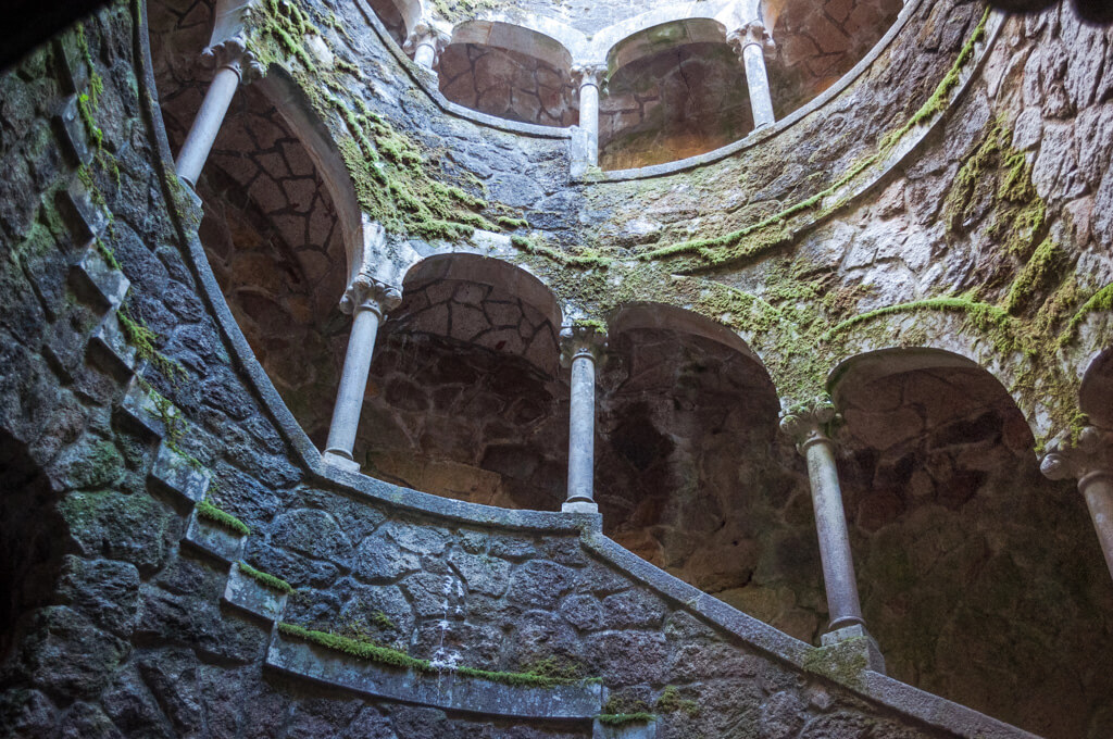 A view of the inside of the Initiation Well at Quinta da Regaleira showing a colonnaded walkway and mossy stone. Depositphotos. 