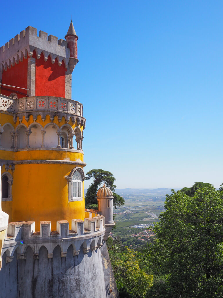 Red and yellow turrets and towers at Pena Palace against a pale blue sky. Copyright@2025 NancyRoberts