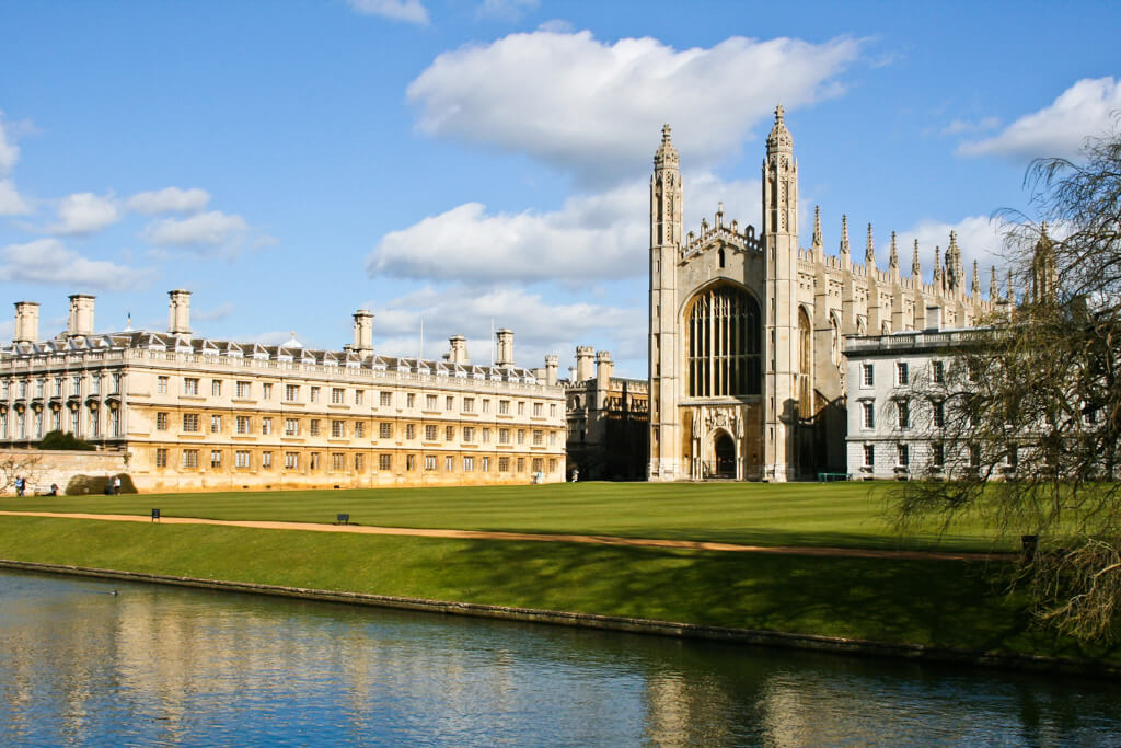 Gothic chapel with two tall spires and college buildings in gold stone overlooking lawns and calm river. DepositPhotos