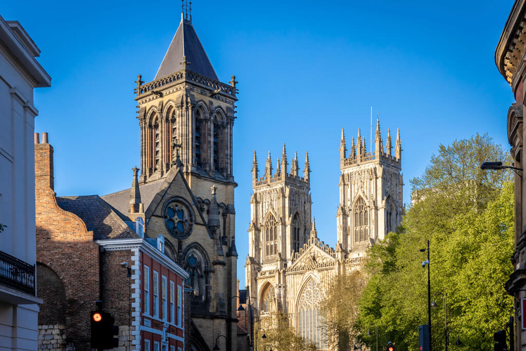 Sunshine on the twin towers of York Minster against a blue sky with another Gothic tower in the foreground. DepositPhotos