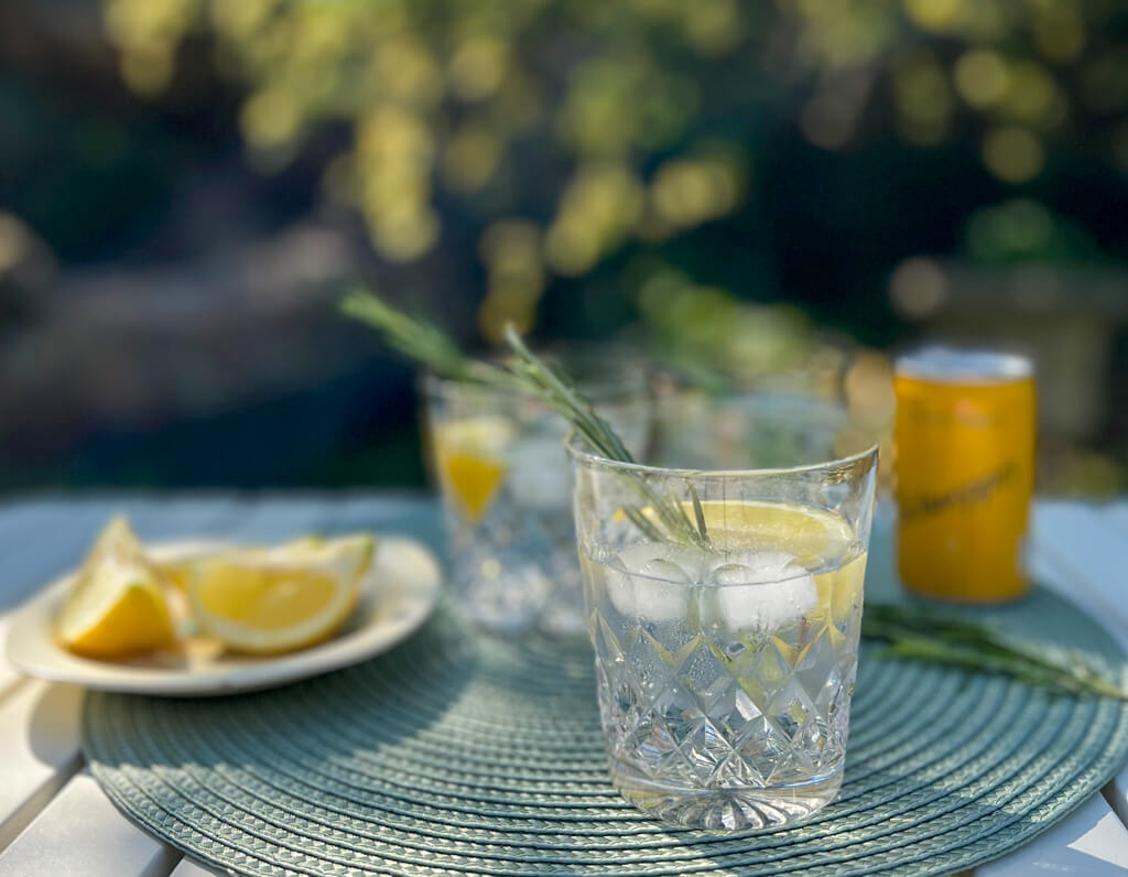 Three tumblers of gin and tonic with sprigs of rosemary and plate of sliced lemon and tin of tonic on table in garden. Copyright@2025 NancyRoberts