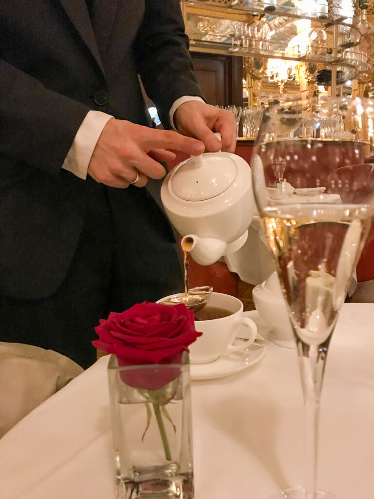 A waiter in suit pours tea with rose and glass of champagne in foreground. Copyright@2025 NancyRoberts