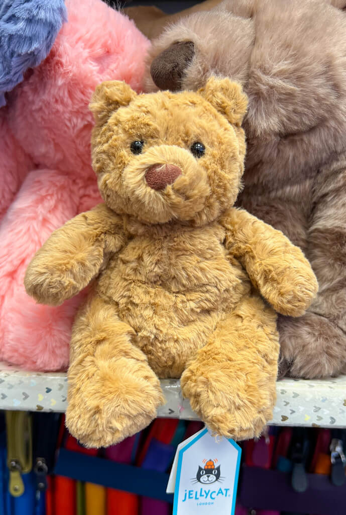 A golden brown teddy bear sitting on a shelf in front of other soft toys. Copyright@2025 NancyRoberts