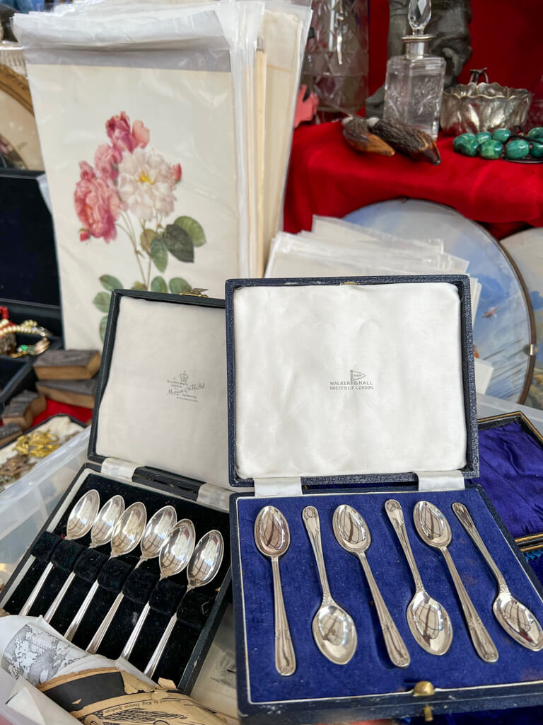 Unique London souveniers: vintage teaspoons in presentation boxes on a market stall with a rose print in background. Copyright@2025 NancyRoberts