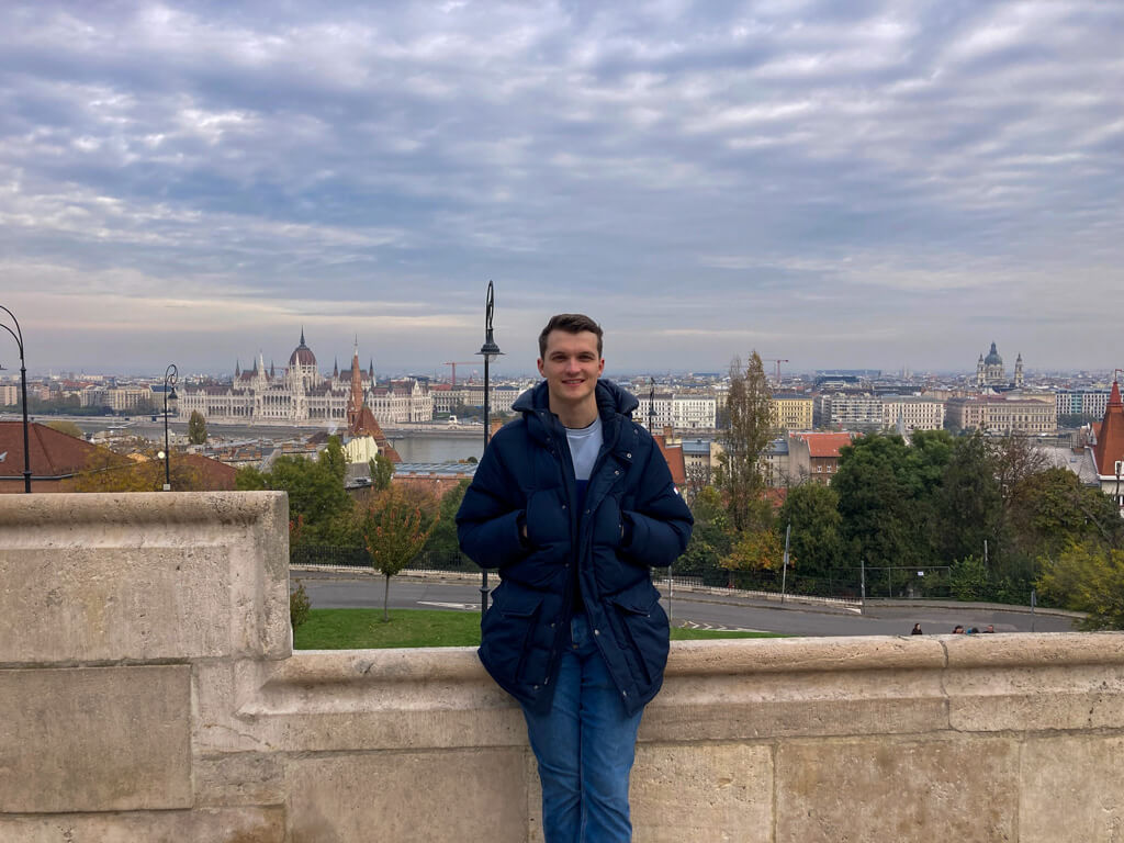 Young man standing on bridge with view of Budapest Parliament building in background. Copyright@2025reserved to photographer via MapandFamily