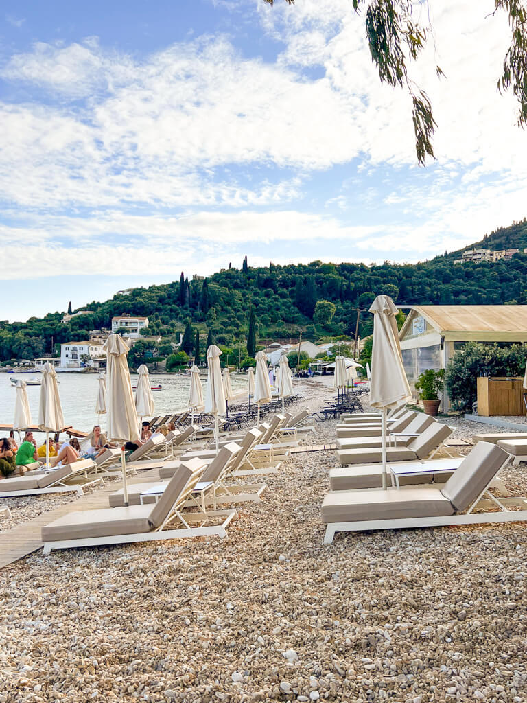 Beach loungers and umbrellas in rows on the sloping pebbly beach of Kalami bay. Copyright@2025 NancyRoberts