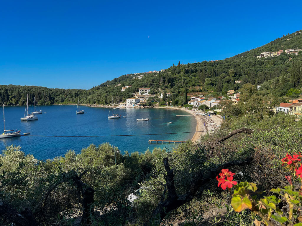 Kalami bay from a path leading to the village, with red geraniums in the foreground and yachts moored in the peaceful blue bay. Copyright@2025 NancyRoberts