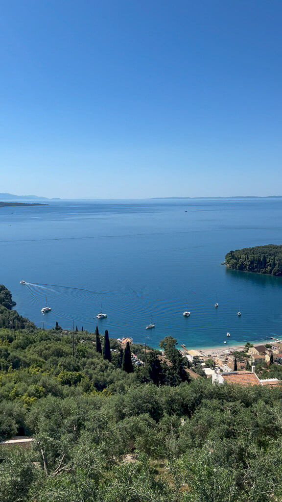 Luxury Corfu villa view over Kalami bay showing forested slopes and low buildings. Copyright@2025NancyRoberts