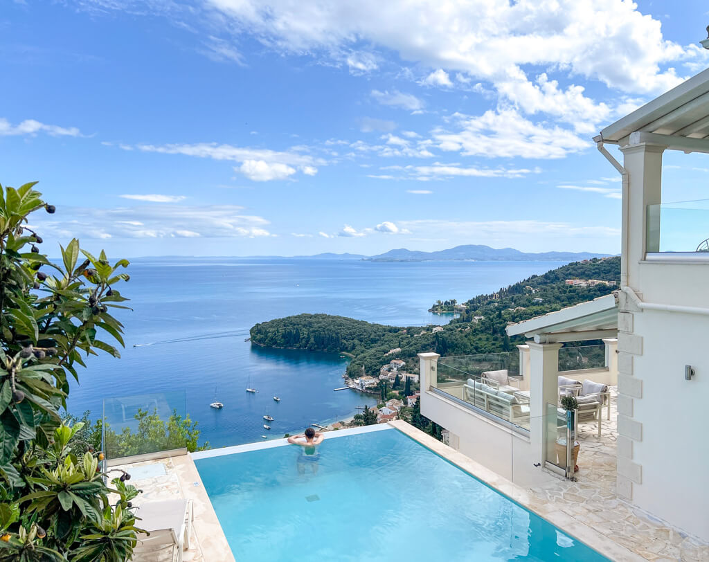 Young man leaning on edge of infinity pool looking out over blue sea of Kalami Bay, Corfu. Copyright@2025 NancyRoberts