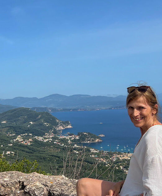 The author, Nancy Roberts, sitting on the ramparts of Anthoussa castle with the coastline and Parga in the distance. Copyright@2025 NancyRoberts