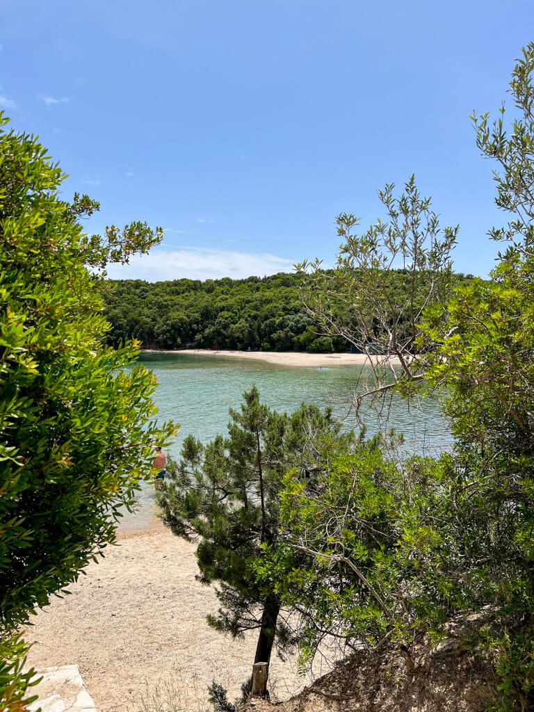 A view framed by shrubs of Bella Vraka and the spit of sand that leads across to the islet. Copyright@2025 NancyRoberts