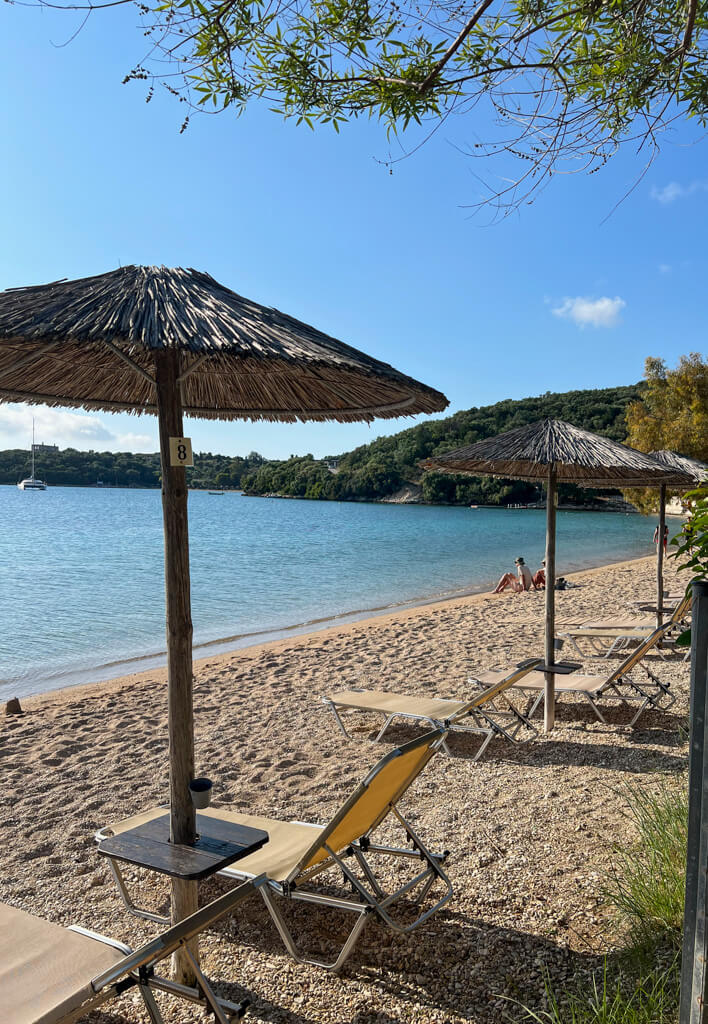 Galikos Molos beach in Syvota, Greece with a line of sunbeds and natural parasols. Copyright@2025 NancyRoberts
