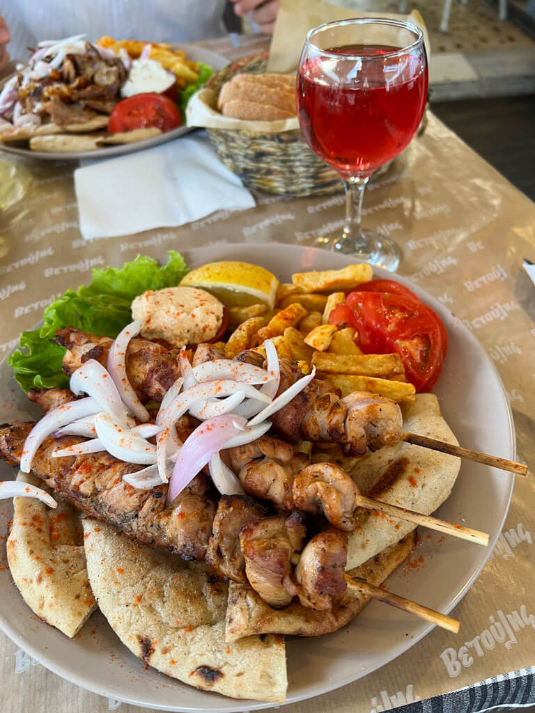 A plate of chicken soulaki topped with onions and served with pitta, tomatoes, chips and lemon. Copyright@2025 NancyRoberts