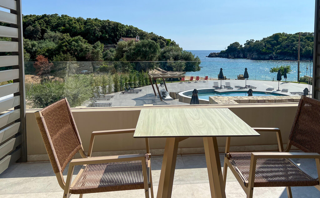 View from balcony at Arete Lifestyle Hotel, Syvota Greece. Modern table and chairs on balcony which looks out to pool, beach and islet. Copyright@2025 NancyRoberts