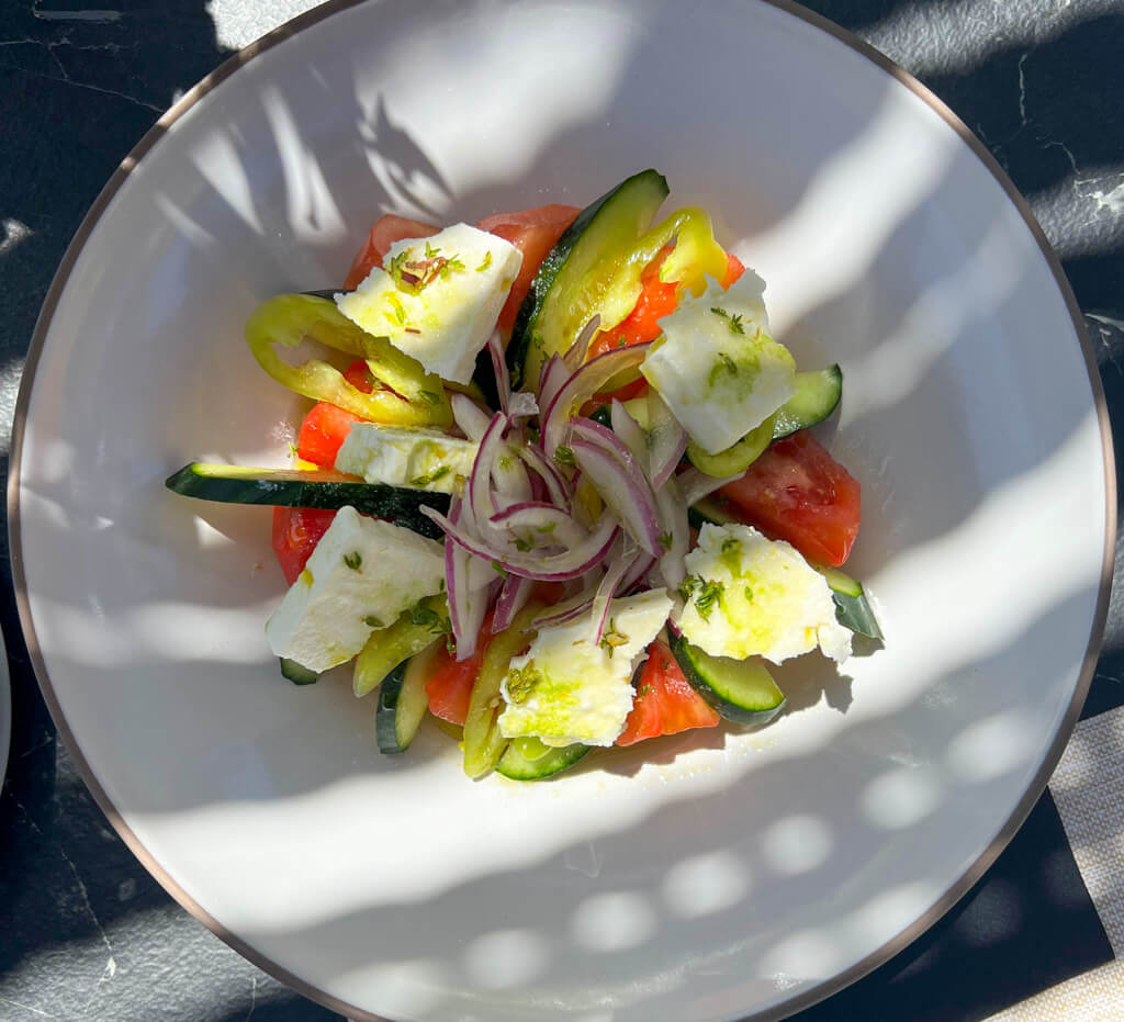 A Greek salad of feta, tomato and onion with a herby dressing in sun dappled shade. Copyright@2025 NancyRoberts