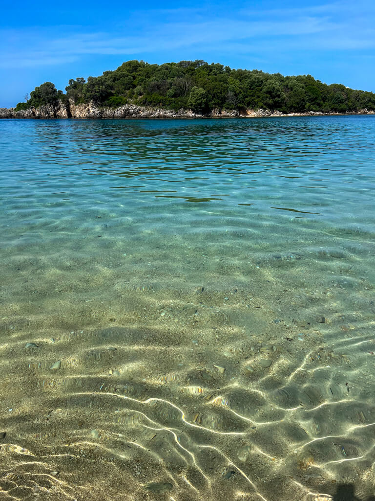 Clear calm turquoise water in the bay at Agia Paraskevi beach. A small wooded islet stands in the middle of the bay. Copyright@2025 NancyRoberts