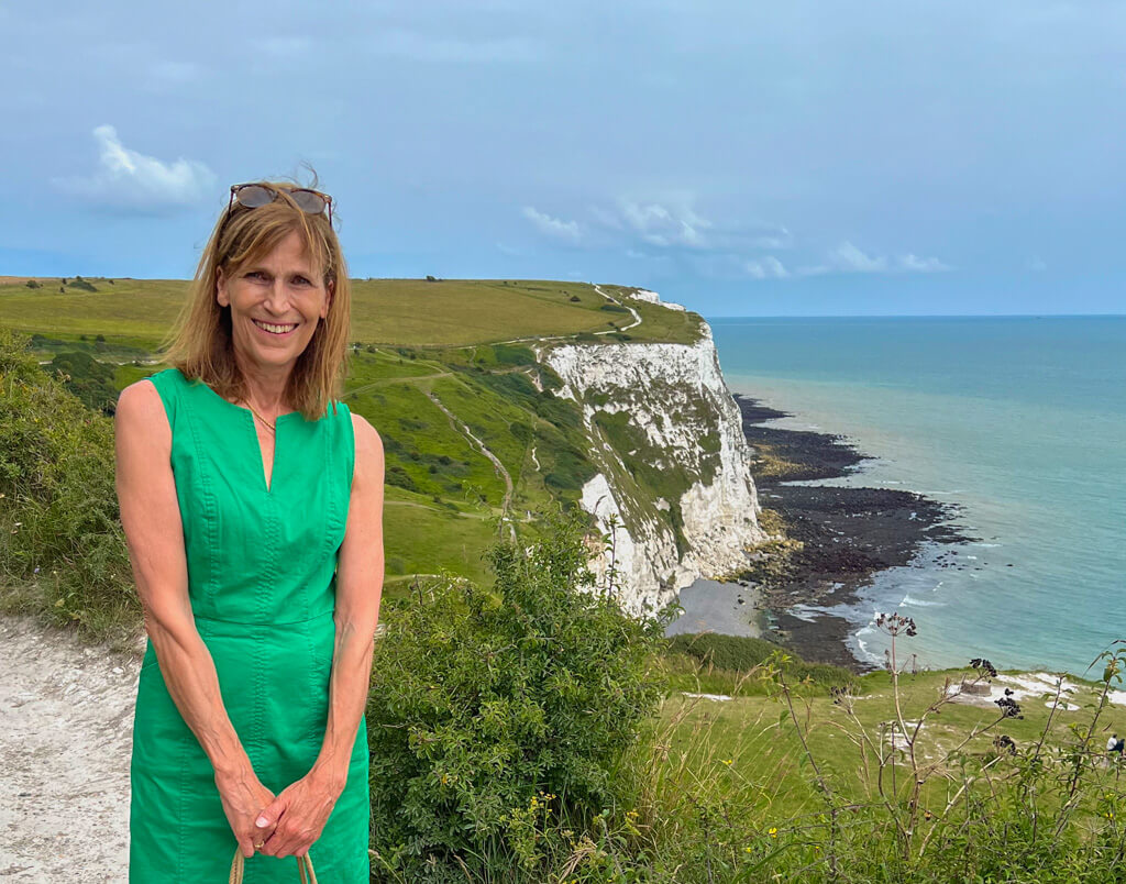 The author, Nancy Roberts, in a green dress, standing on the clifftops at Dover with the White Cliffs in the background. Copyright@2025 Nancy Roberts