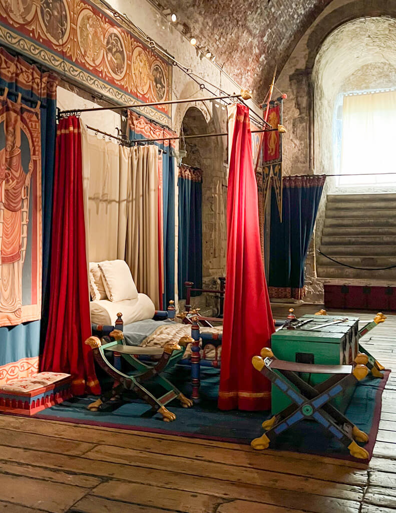 Richly coloured red bed hangings, a green chest and blue and gold stools in a re-creation of a medieval aristocrat's bed in Dover Castle.  Copyright@2025 Nancy Roberts